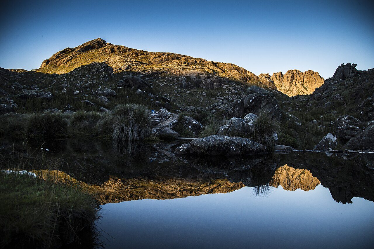Ao entardecer, um espelho d'água reflete encostas de capim e blocos de granito; ao fundo, à direita, o paredão recortado das Agulhas Negras recebe a última luz do sol.