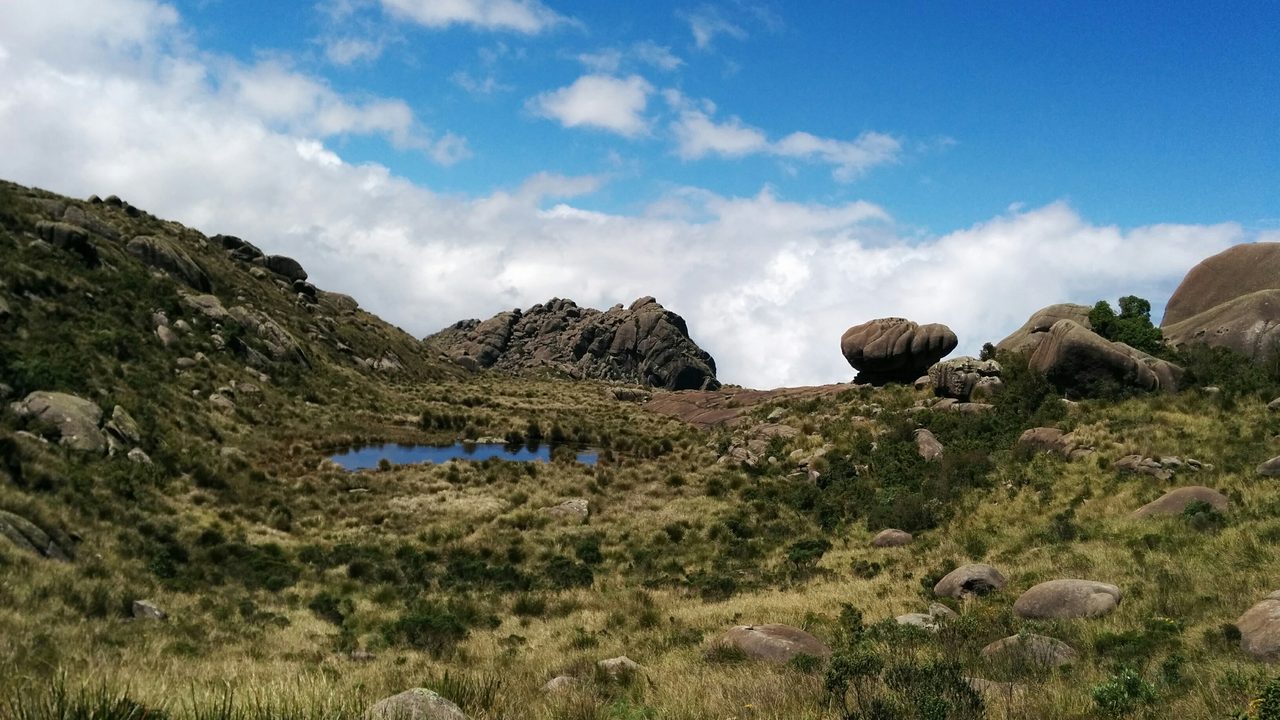 Campo de altitude com touceiras de capim e uma poça d'água rasa refletindo o céu; espalhados pelo terreno, blocos de granito arredondados, um deles equilibrado em forma de cogumelo, e ao fundo um paredão de rocha sob céu de nuvens.