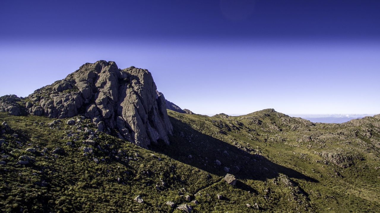 Ao amanhecer, um bloco de rocha fraturado sobe acima de encostas de capim baixo salpicadas de pedras; à direita o terreno desce em lombadas até um horizonte de serras, sob céu limpo.