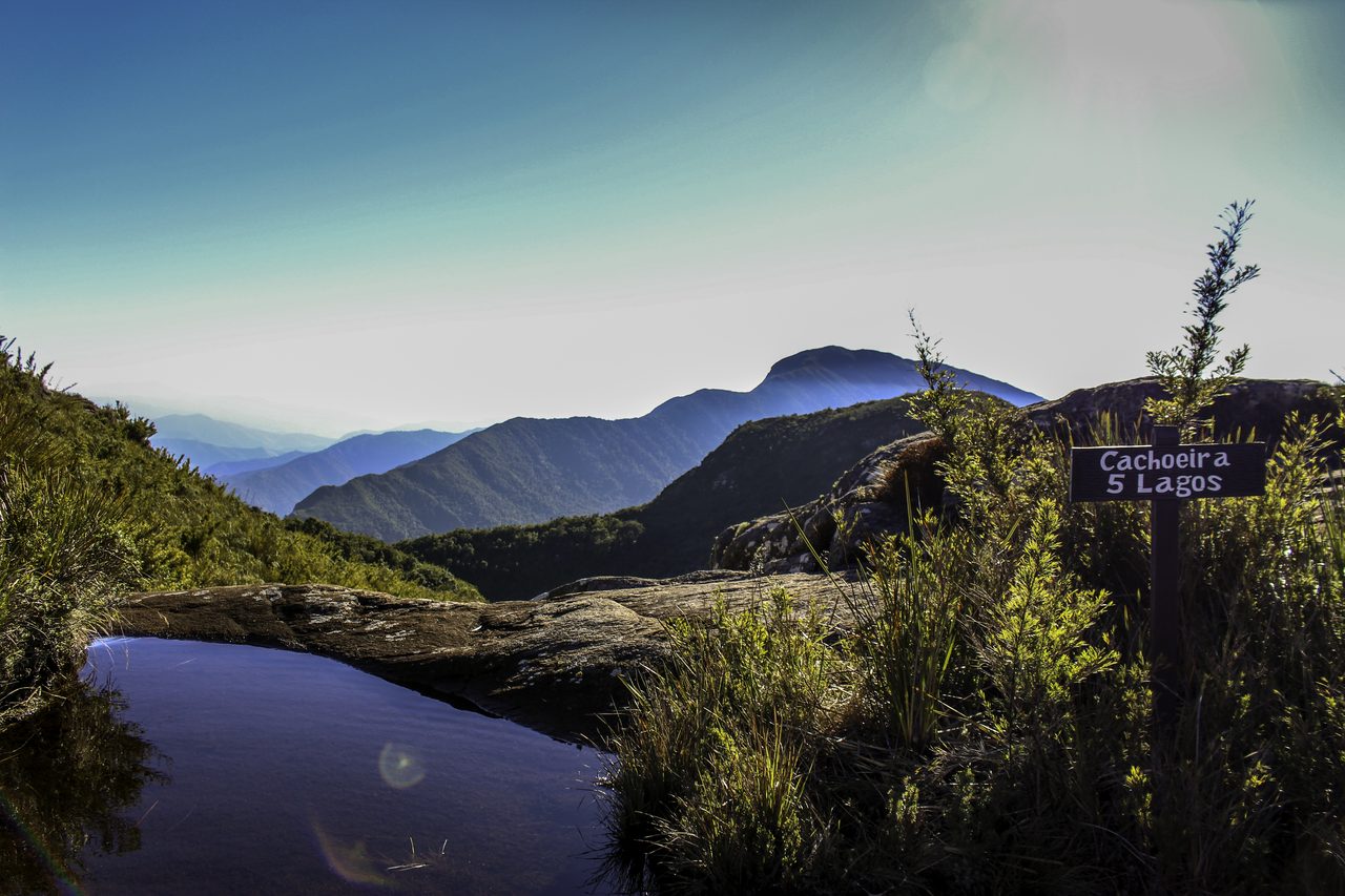 Uma lâmina de água parada sobre laje de pedra, na beira de um patamar de campo de altitude; à direita, uma placa de madeira escrita "Cachoeira 5 Lagos" entre touceiras de capim, e ao fundo serras azuladas em camadas até o horizonte.