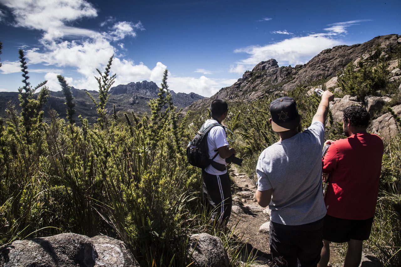 Trilha de terra clara subindo por encosta de capim baixo salpicada de pedras; à esquerda o terreno cai para um vale, e ao fundo cadeias de montanhas se sobrepõem até o horizonte, sob nuvens.