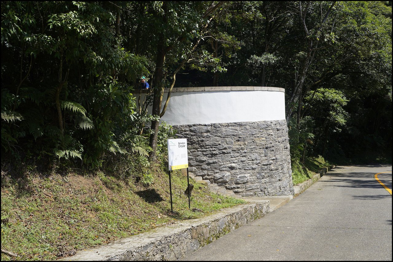 Mirante de pedra em formato circular à beira da estrada asfaltada, cercado pela mata, com uma placa informativa ao lado