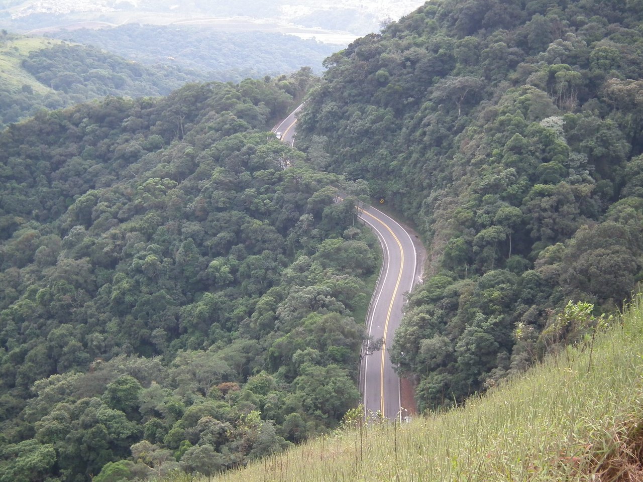 Estrada asfaltada sinuosa serpenteando entre morros cobertos de mata, vista do alto