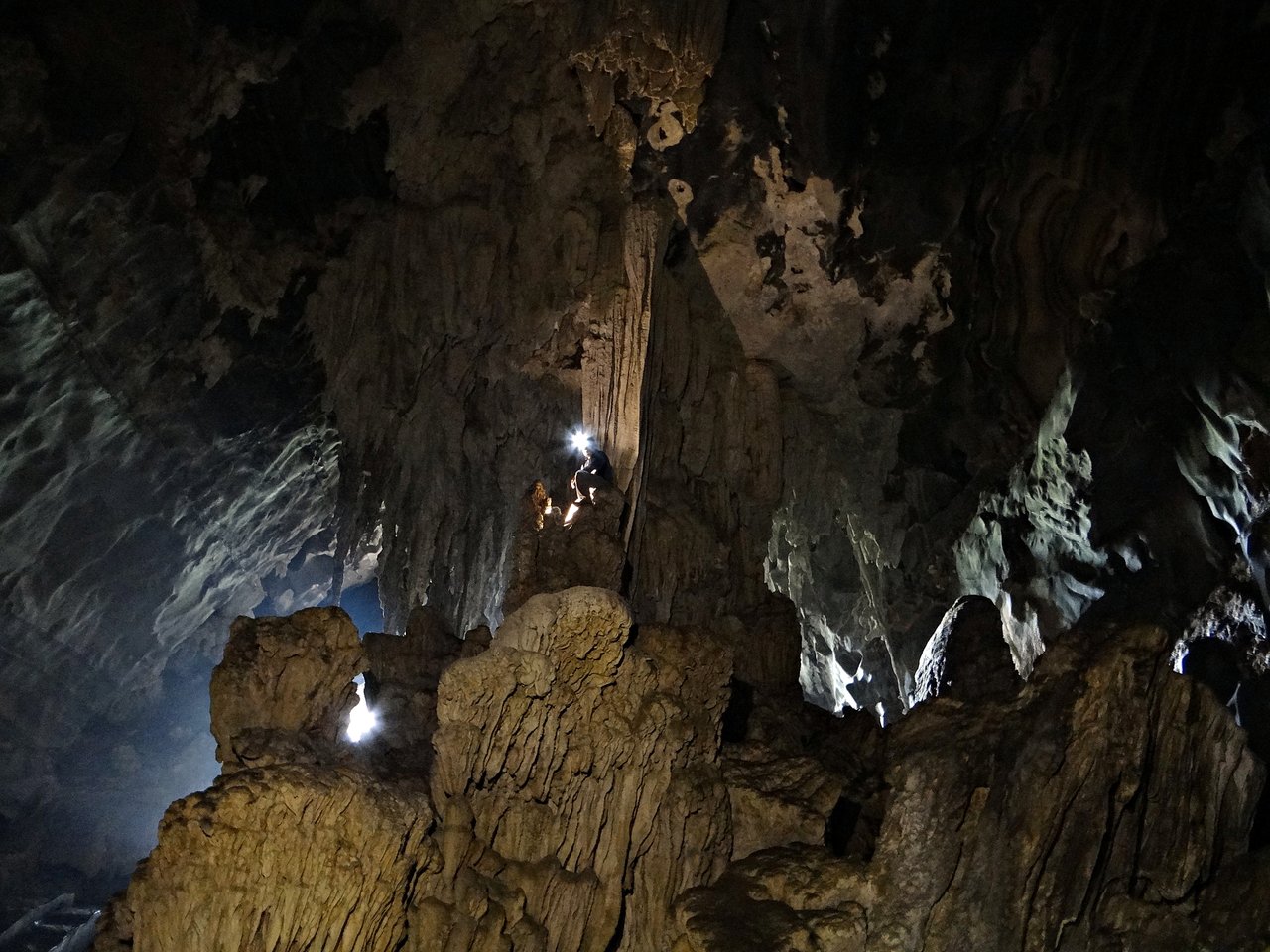 Interior de caverna com estalactites e formações de rocha clara, iluminado por lanternas de visitantes ao fundo