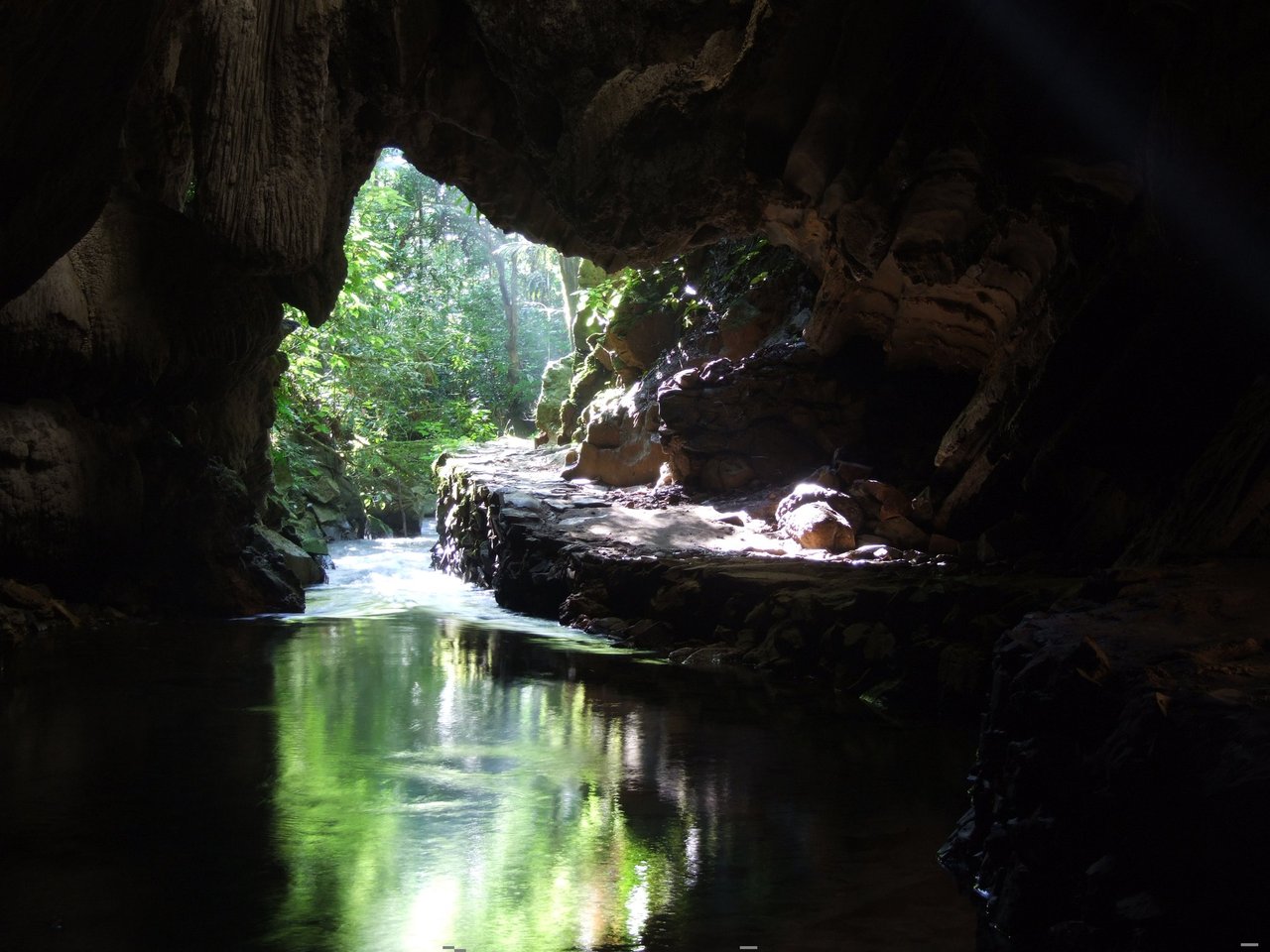 Boca de caverna vista de dentro, com um rio de águas esverdeadas correndo sob a rocha rumo à luz e à mata