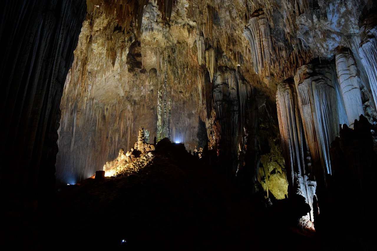 Grande salão de caverna com estalactites e colunas de rocha clara, com pontos de luz de lanternas ao fundo