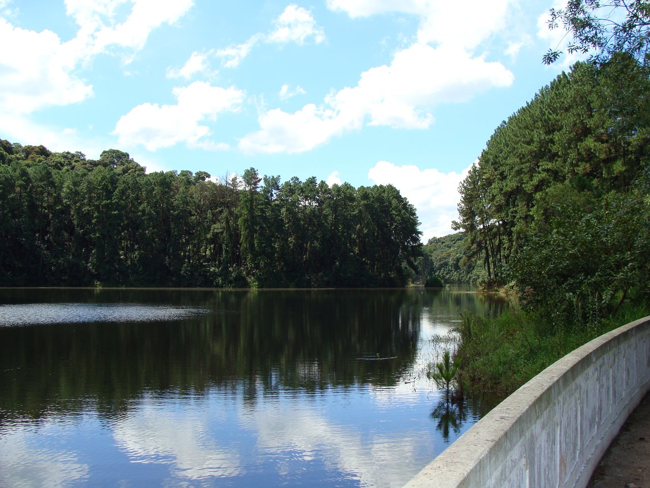 Represa do Cabuçu, com a água espelhando as árvores da margem e uma mureta de concreto à direita, sob céu com nuvens